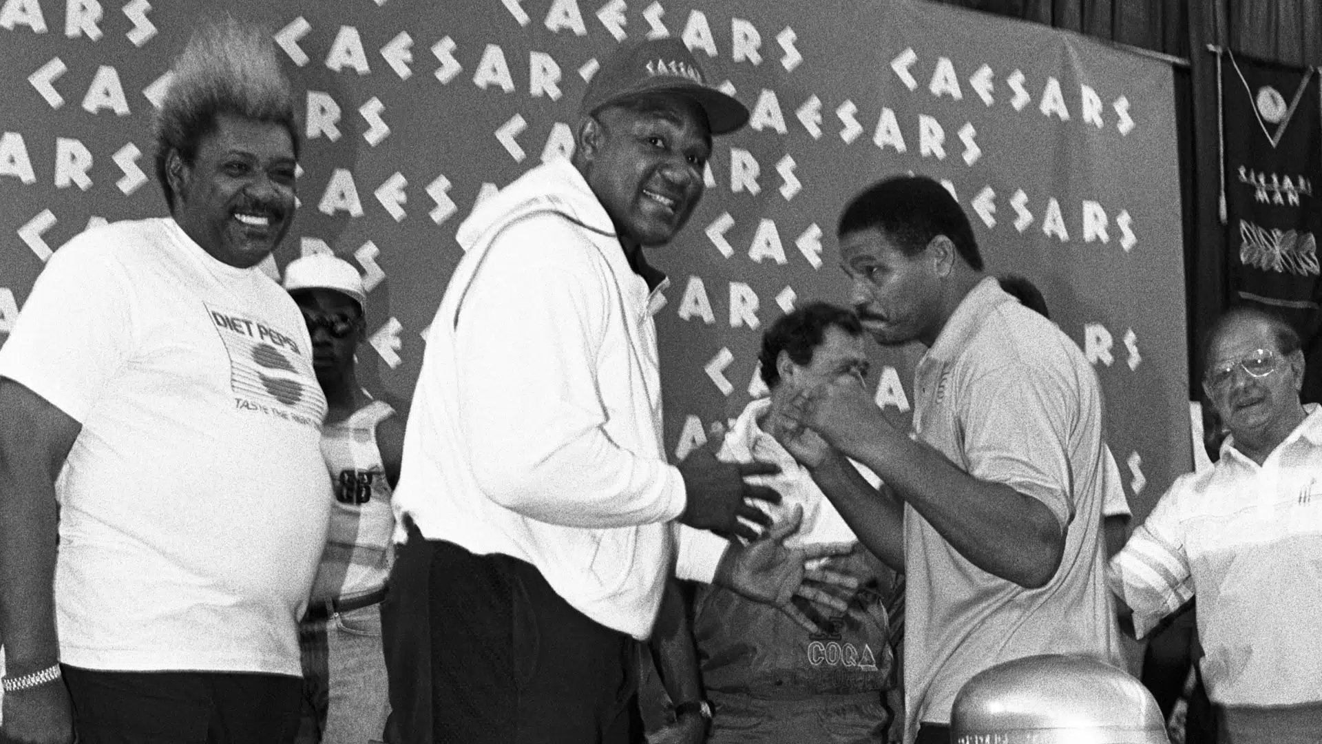 Encarada entre George Foreman e Maguila, com Angelo Dundee ao fundo, no lado direito da imagem 